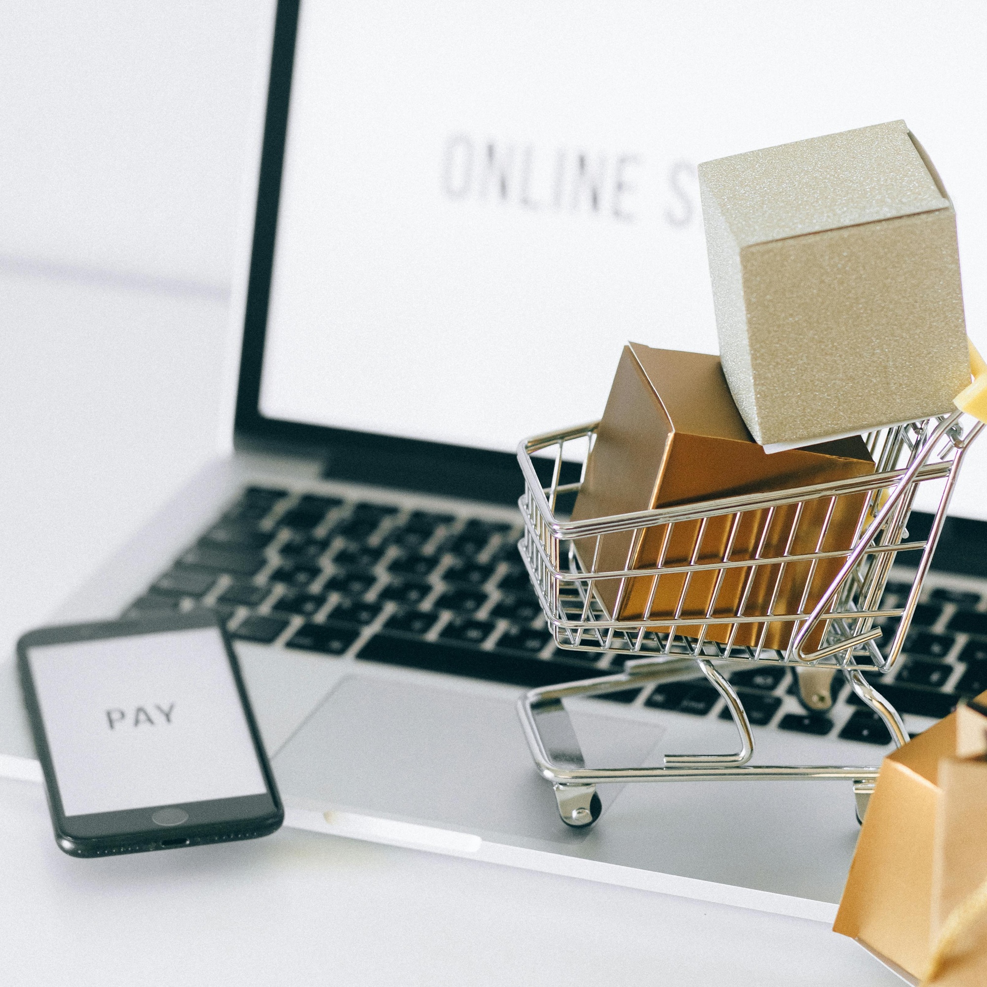 Laptop displaying an online store with a miniature shopping cart full of boxes and a smartphone showing PAY on screen