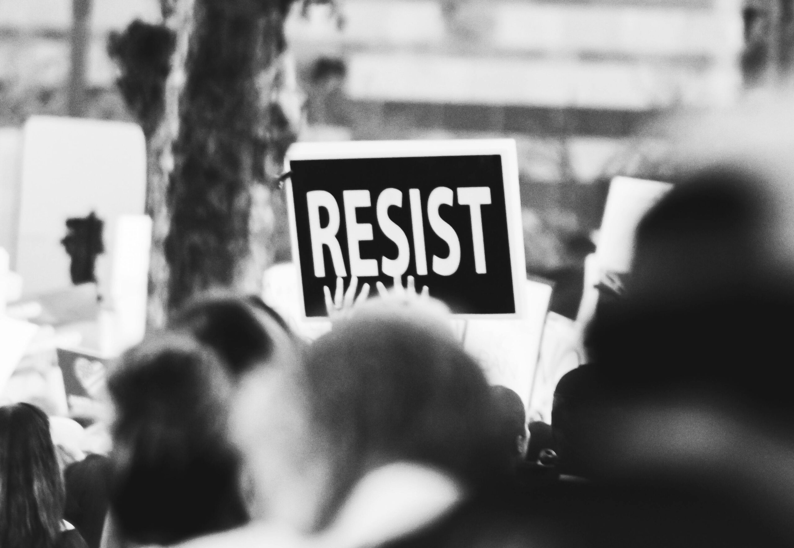 Black and white photograph of a crowd at a protest with someone holding a sign that reads RESIST