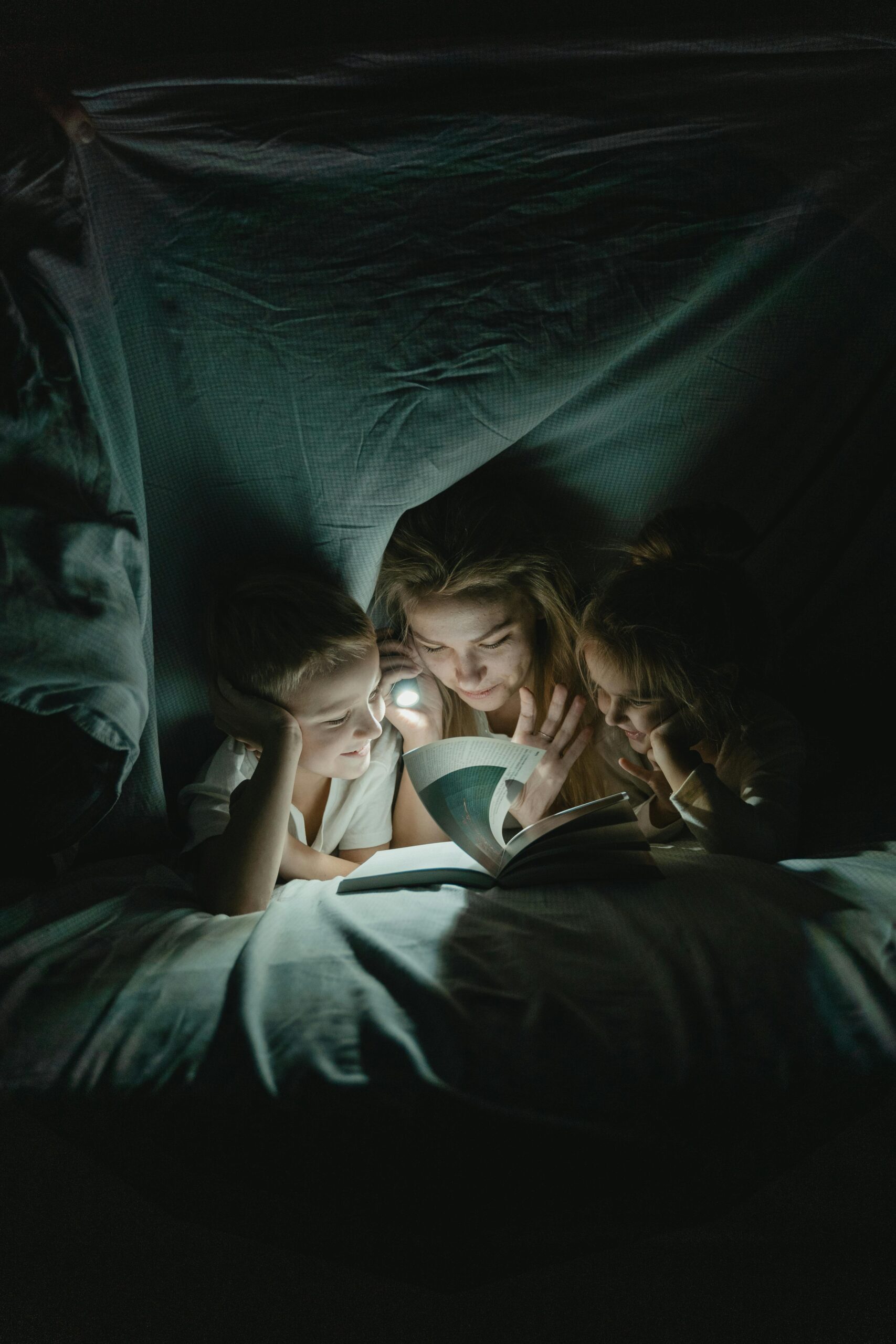 Mother and two children reading a book together under a blanket by flashlight in the dark